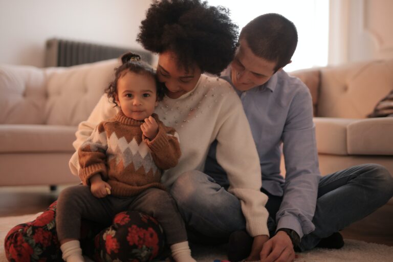 Mama Sarah, Papa David und kleine Tochter Emma im Wohnzimmer lachend, Emma schaut süß in die Kamera
