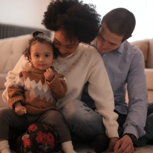 Mama Sarah, Papa David und kleine Tochter Emma im Wohnzimmer lachend, Emma schaut süß in die Kamera