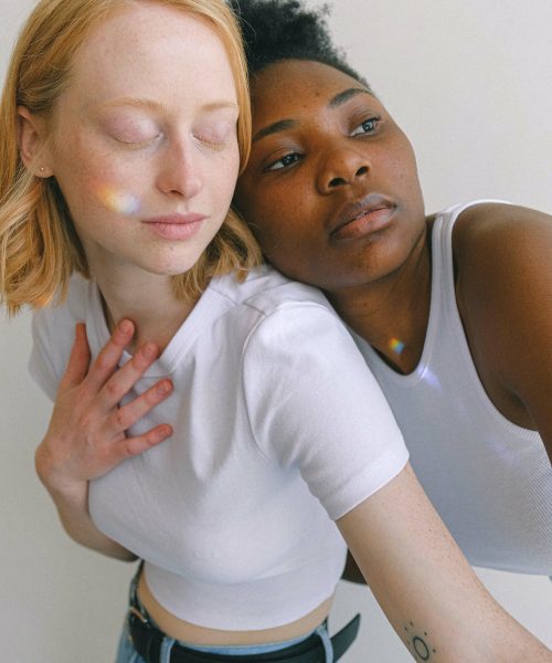 Ava lehnt sich von hinten auf Mayas Schulter, Regenbogen Lichtspiel über Gesichter der beiden Frauen